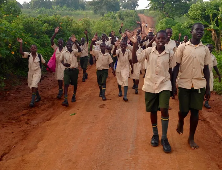 Group of schoolchildren in uniforms walking and waving along a dirt road surrounded by greenery