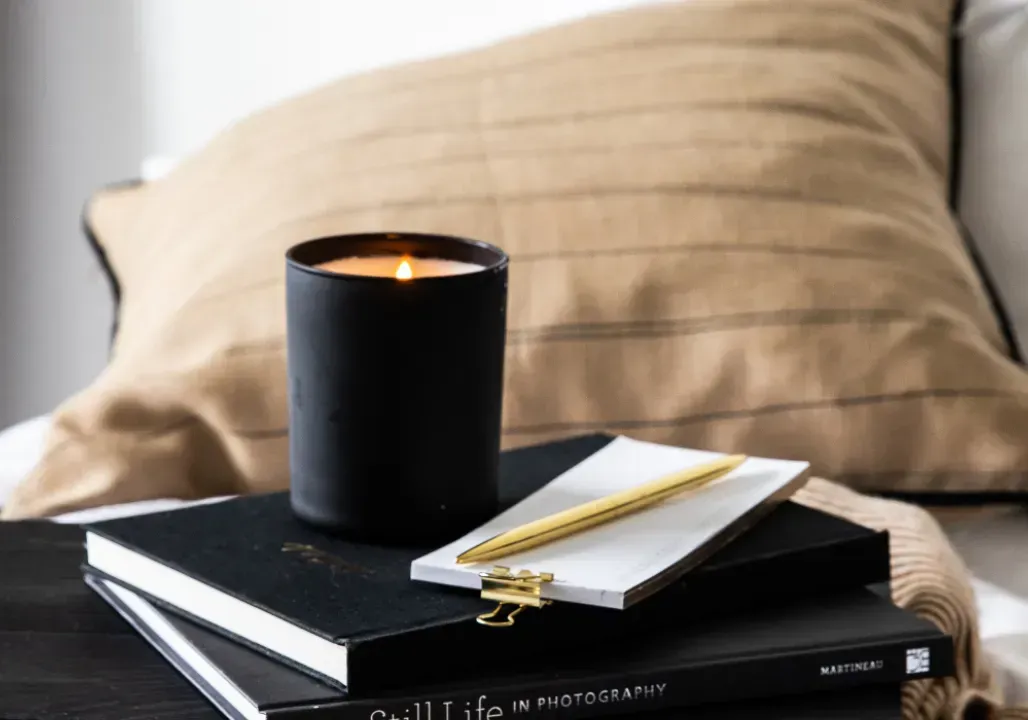 Black candle, gold pen, and notebooks on a cozy bed with a tan pillow, symbolizing reflection, clarity, and soulful business planning
