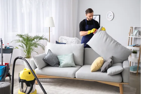 Professional cleaner in a blue apron and yellow gloves lifts a cushion while tidying a well-decorated living room with cleaning supplies and a vacuum nearby