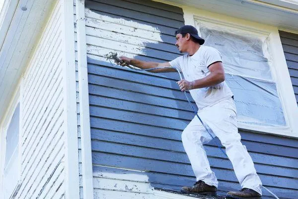 A man in white work clothes is painting the exterior of a house. He is using a paint sprayer to apply a fresh coat of paint to the siding, standing on a ladder for better reach.