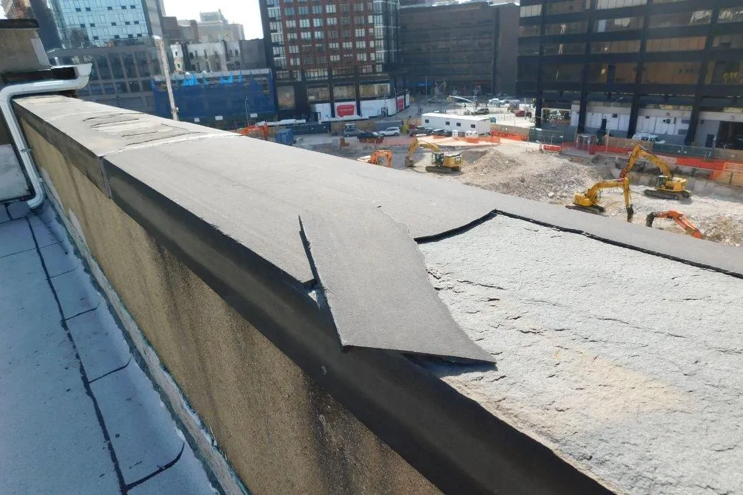 A close-up view of a parapet wall with visible weather damage, including peeling and cracked material on the top surface. The background shows a construction site with machinery, emphasizing the urban setting. The image highlights the effects of weather on the parapet's durability, making it relevant for discussions about maintenance and protection in harsh climates like New York City.