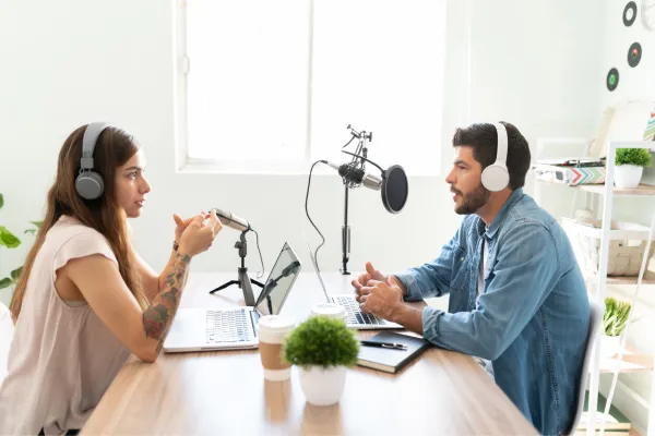 Two people siting across from each other, they are both wearing headphones and there is a podcast microphone sitting between them