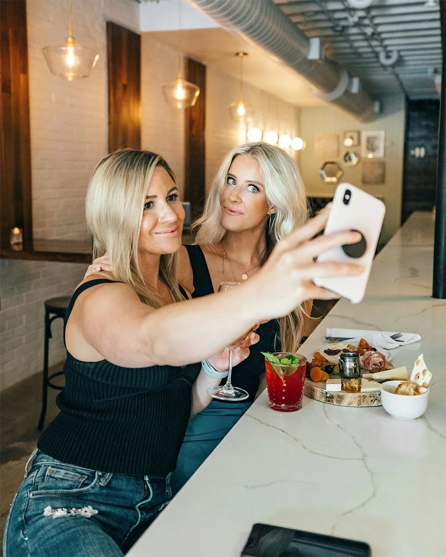 "Two stylish women taking a selfie at a modern bar, enjoying cocktails and a charcuterie board, embracing friendship and fun moments together.