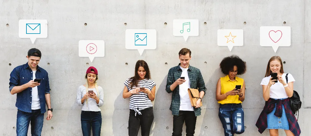 Six diverse young adults standing against a wall, each engaged with their smartphones. Above their heads are speech bubbles with icons representing different forms of digital content and marketing: email, video, photo, music, star rating, and heart. The image highlights the use of various digital marketing channels.