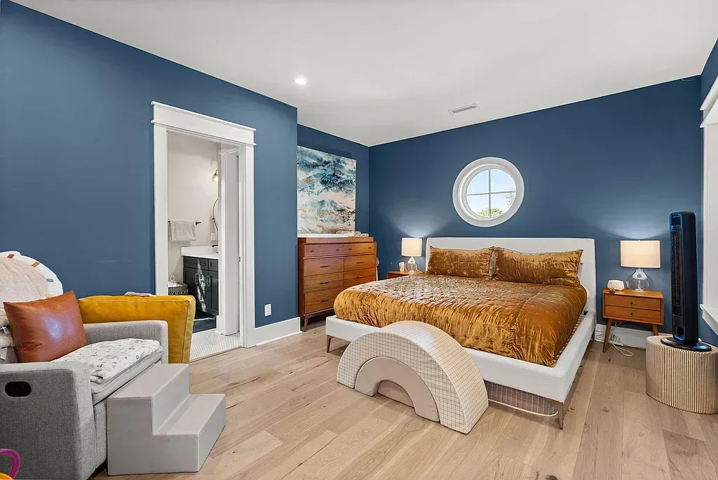 Modern luxury bedroom with bold blue walls, round window, wood furniture, and gold bedding in Oklahoma home
