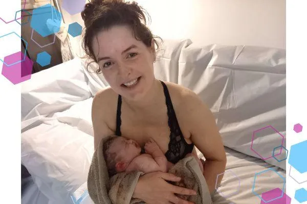 Rachel sitting on the sheet covered sofa holding her newborn baby in her arms. Rachel is smiling at the camera after her home birth in Welwyn.