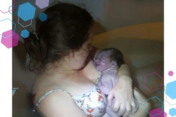 Jilly sitting in a birth pool cuddling her brand new baby daughter. Both mum and baby are looking at each other. The room is dark and the baby is so new she looks grey or purple.