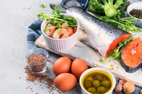 The image displays a variety of healthy foods, ideal for a balanced diet. It includes a large, fresh salmon fillet with its silvery skin visible, alongside a smaller portion of sliced smoked salmon. Adjacent to the fish, there's a bowl of peeled shrimp, ready to eat. The scene also features a small glass bowl of flaxseeds, a significant source of Omega-3 fatty acids, and a bowl of green olives. Additionally, there are three whole, brown eggs and a collection of green vegetables and herbs in the background, emphasizing a focus on nutritious, wholesome ingredients.