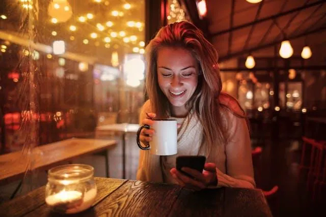 Smiling woman sitting in a cosy café at night, holding a mug and looking at her phone, symbolising the freedom and clarity teachers can find after leaving the classroom.