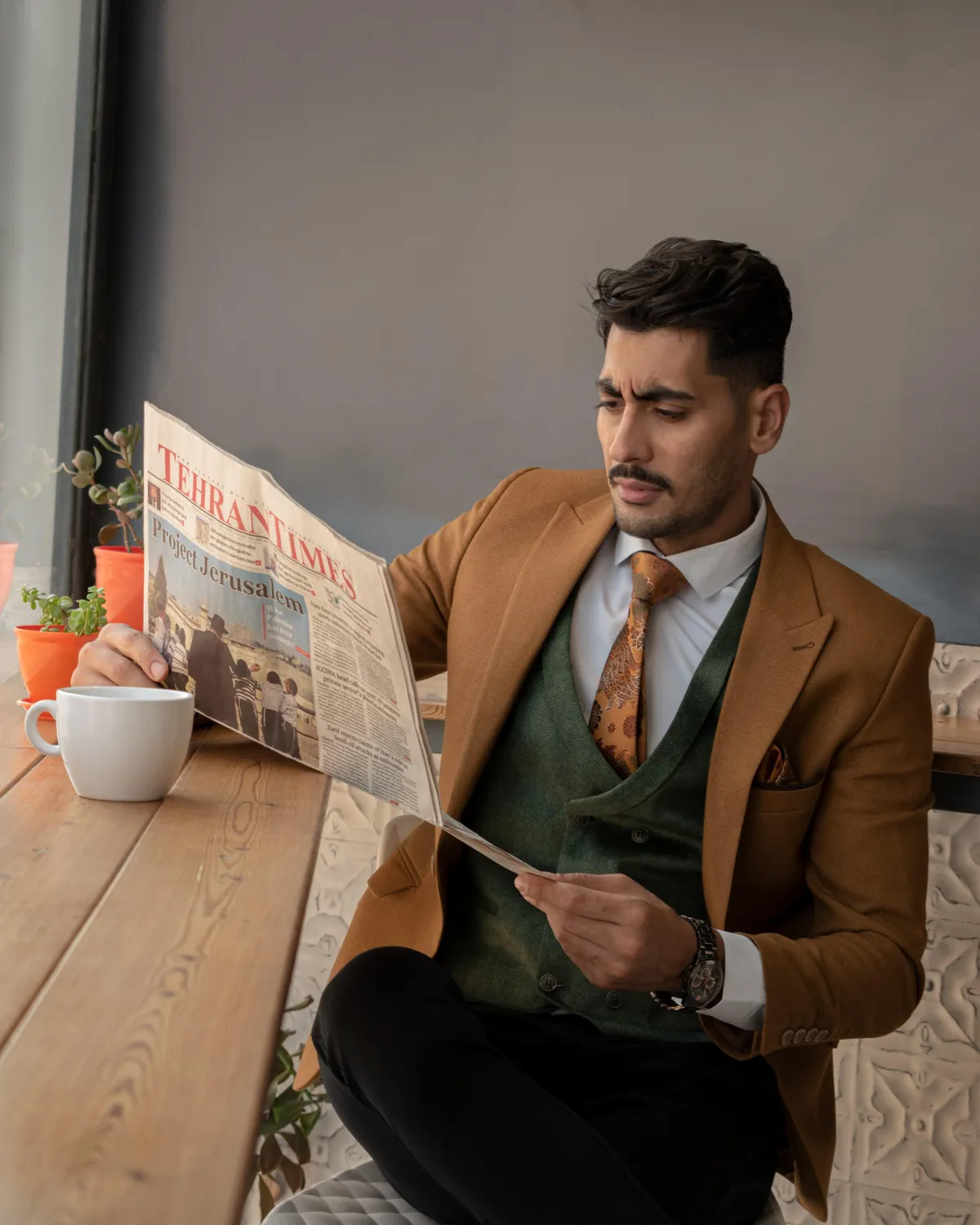 A man in a custom brown blazer, green waistcoat, black pants reading a newspaper