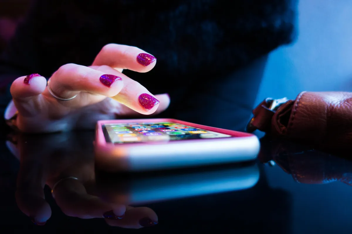 A close-up photo of an iPhone laying on a table as a woman is about to click an app on the Phone.