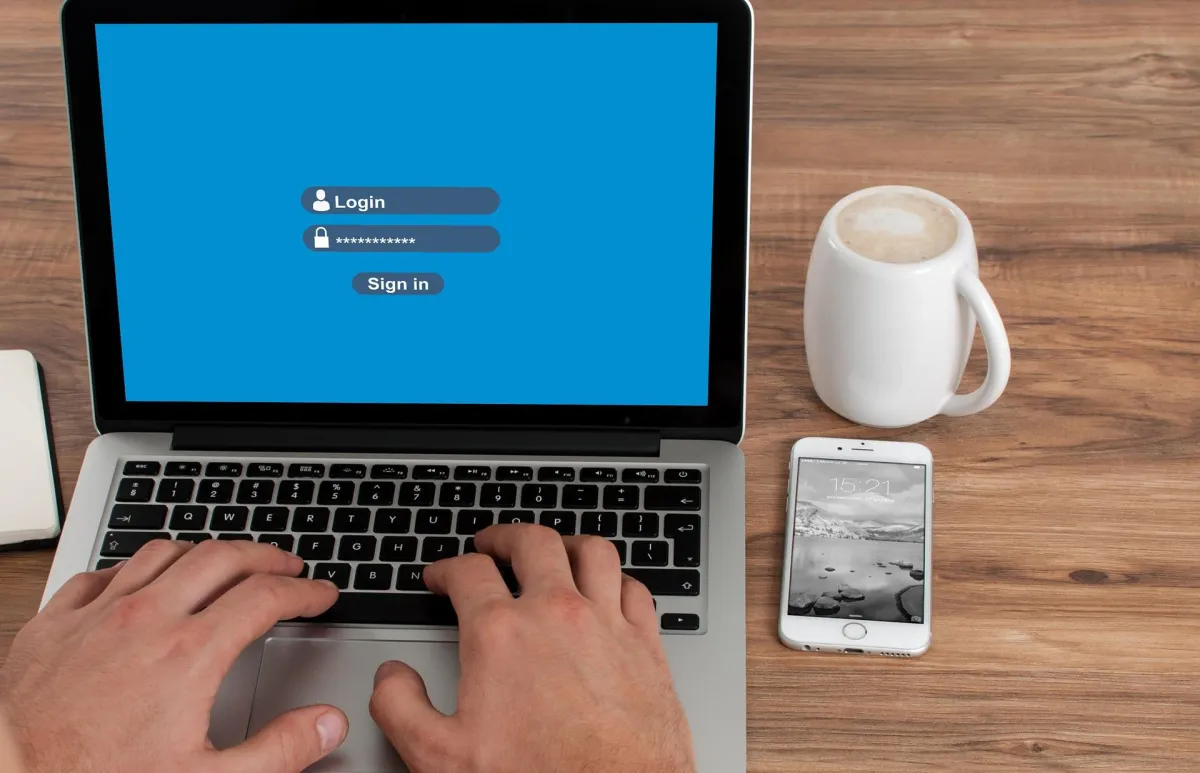A person logging into their laptop with an iPhone and a coffee beside them.