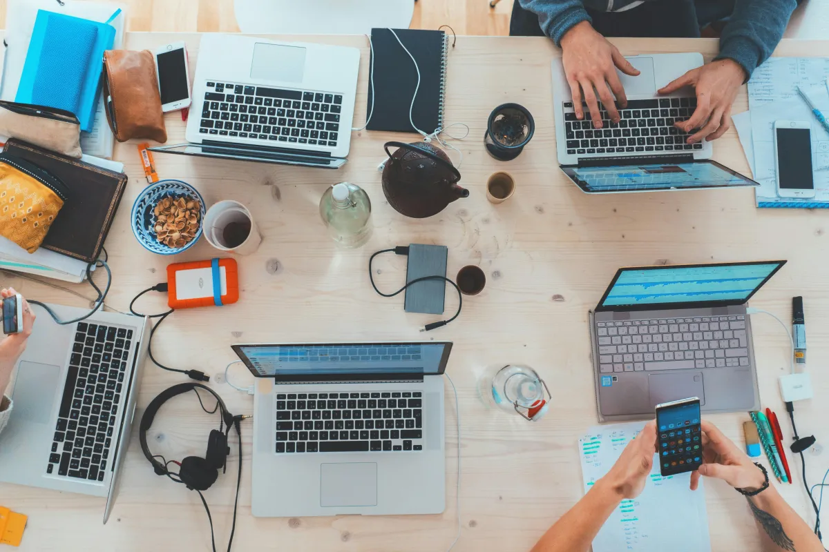 people sitting down near a table with assorted laptops, computers, phones, chargers and headphones.
