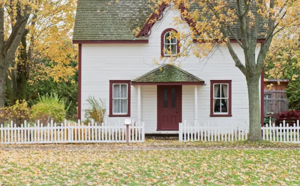 Charming white house with a red door and picket fence surrounded by autumn trees, symbolizing warmth, stability, and thoughtful decision-making about where to live