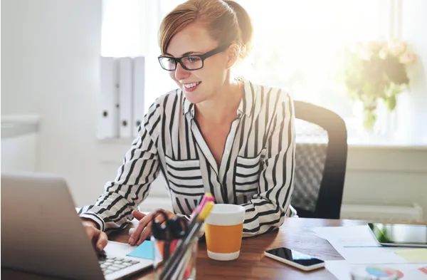 Smiling woman working at her laptop with notes and coffee on the desk, representing productivity, focus, and SMART goal setting.
