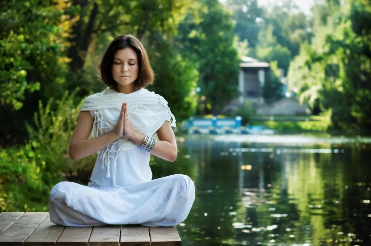 “Woman meditating peacefully by a lake surrounded by greenery, symbolizing mindfulness, inner awareness, and overcoming negative self-talk