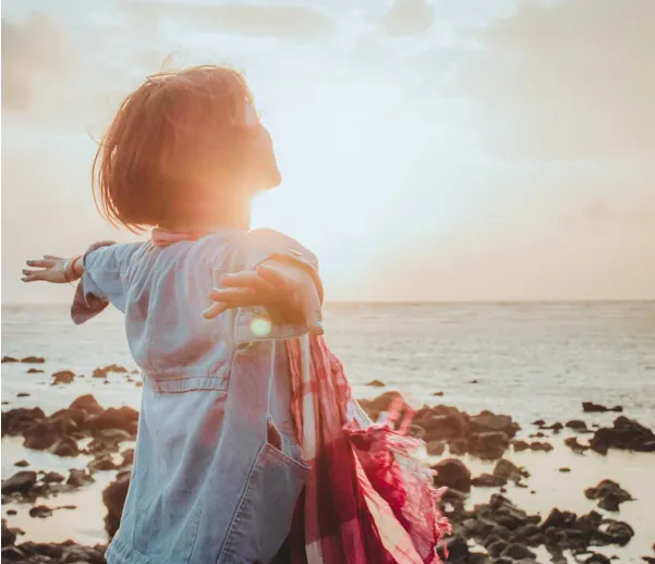 Joyful woman standing by the ocean with open arms at sunrise, symbolizing balance, reflection, and fulfillment across all life areas.