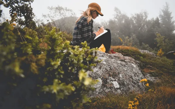 Woman sitting on a rock in nature, writing in a journal surrounded by greenery and mist, symbolizing reflection, clarity, and personal growth planning