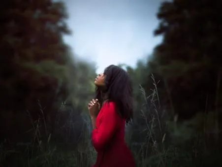 Woman in nature wearing a red dress, gazing upward with hands held together, expressing gratitude and inner peace