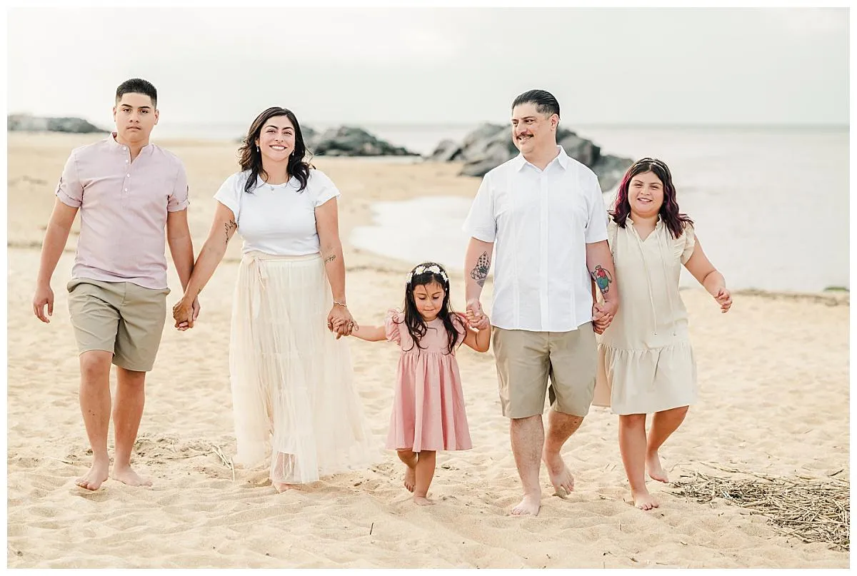 A family of five walks barefoot along the beach at Fort Story, holding hands. They are dressed in whites, khakis, and pinks.