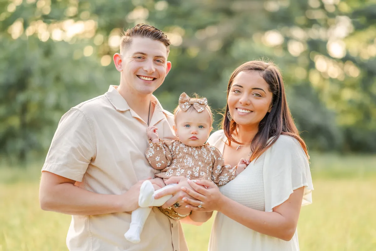Family family of three in a grassy field. Dressed in whites, the parents hold their baby daughter, who is in a brown floral romper, between.