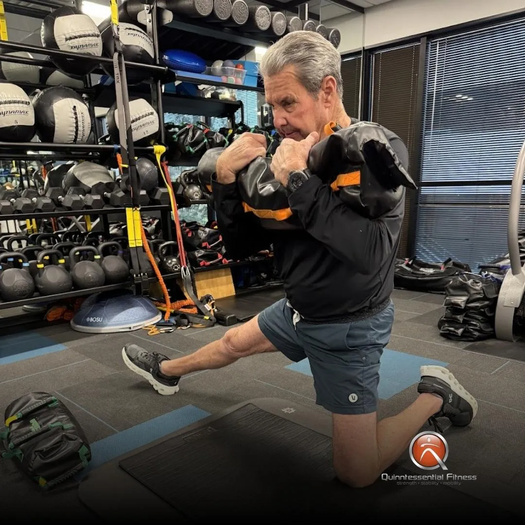 An older adult male performs a loaded split squat with a sandbag at Quinntessential Fitness, focusing on strength and balance to support an active lifestyle."