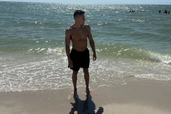 A man stands on the beach with his arms raised, enjoying the ocean breeze and the sunny sky.