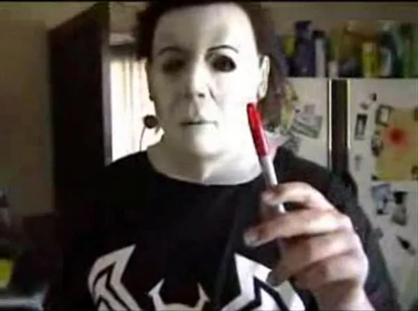 A man with a scary mask on standing in his room with a sharpie in his hand