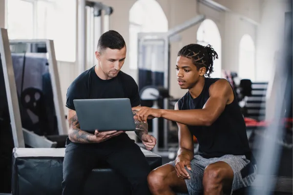 A personal trainer and client discussing progress with a digital tablet showing charts, in a gym setting, symbolizing mentorship and strategic business planning.