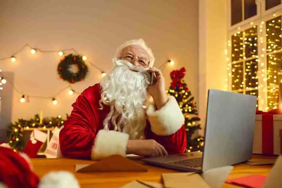 Santa Claus Working on Laptop While Talking by Phone in Room Decorated for Christmas.