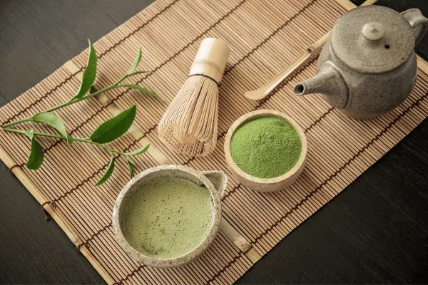 Egy bambusz alátét, rajta balról jobbra: egy tál, benne vízben feloldott matcha por, egy másik tál, benne a matcha porral, egy kancsó, mellettük pedgi egy bambusz ecset és egy ág, néhány levéllel