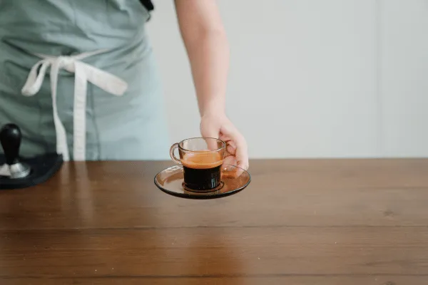 Egy fából készült asztallap, amire egy világoskék kötényt viselő ember épp lerak egy kicsi pohár tiszta fekete kávét, ami egy csészealjon ül, és a bal alsó sarokban egy tömörítő állomás is látható