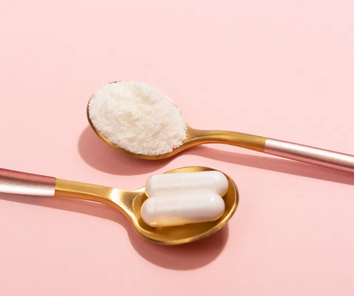 Two golden spoons placed on a soft pink surface, one holding white collagen powder, and the other containing two white collagen capsules. The image emphasises the beauty and simplicity of collagen supplements as part of a wellness routine