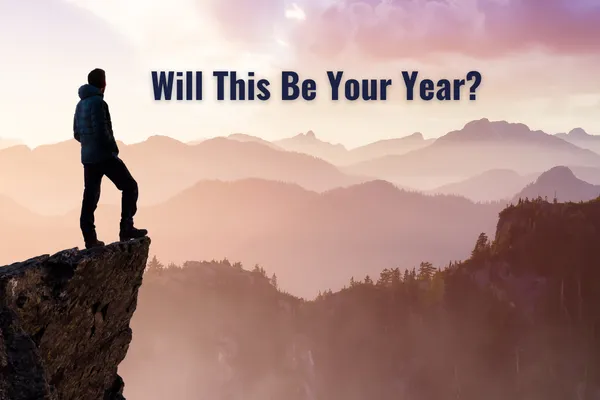 A person stands confidently on the edge of a rocky cliff, overlooking a vast, misty mountain range during sunrise. Soft pink and purple hues fill the sky, symbolizing new beginnings and endless possibilities. Bold text across the sky reads, “Will This Be Your Year?” conveying a sense of reflection and motivation for taking action.
