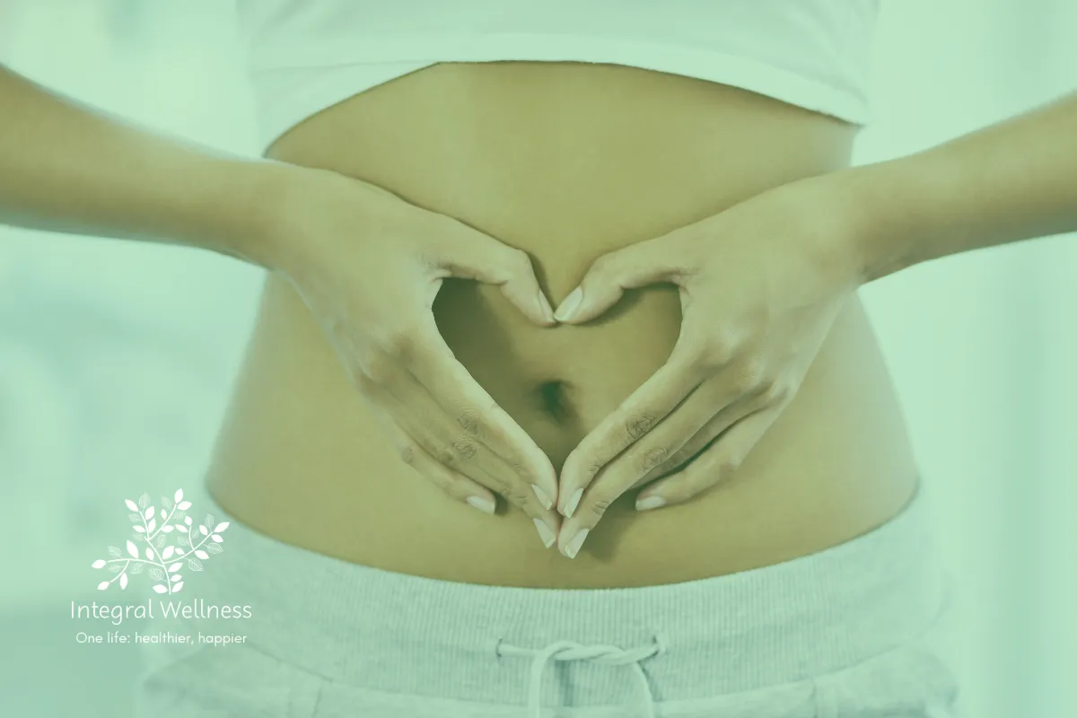 Front view of a woman's belly, wearing white pants and sprots bra, showing the belly button on the center. Her hands forming a heart shape are placed on the belly and around the belly button. The Integral Wellness logo appears in the bottom left corner.