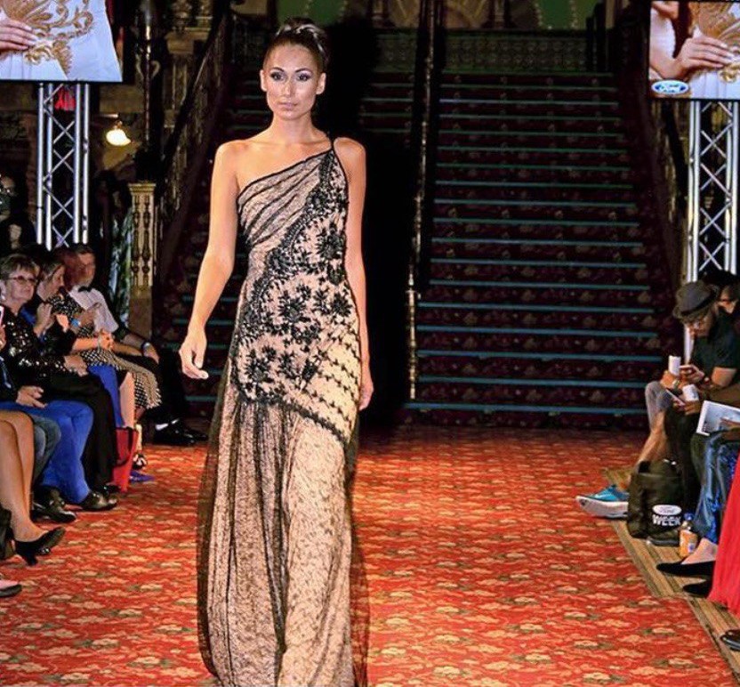 A female fashion model walks down a runway wearing an elegant, one-shoulder floor-length gown. The dress is a light beige or nude color, overlaid with intricate black lace patterns and floral embroidery that drape diagonally across the bodice. She is walking on a red patterned carpet in a grand venue with a large staircase and an audience seated on both sides.
