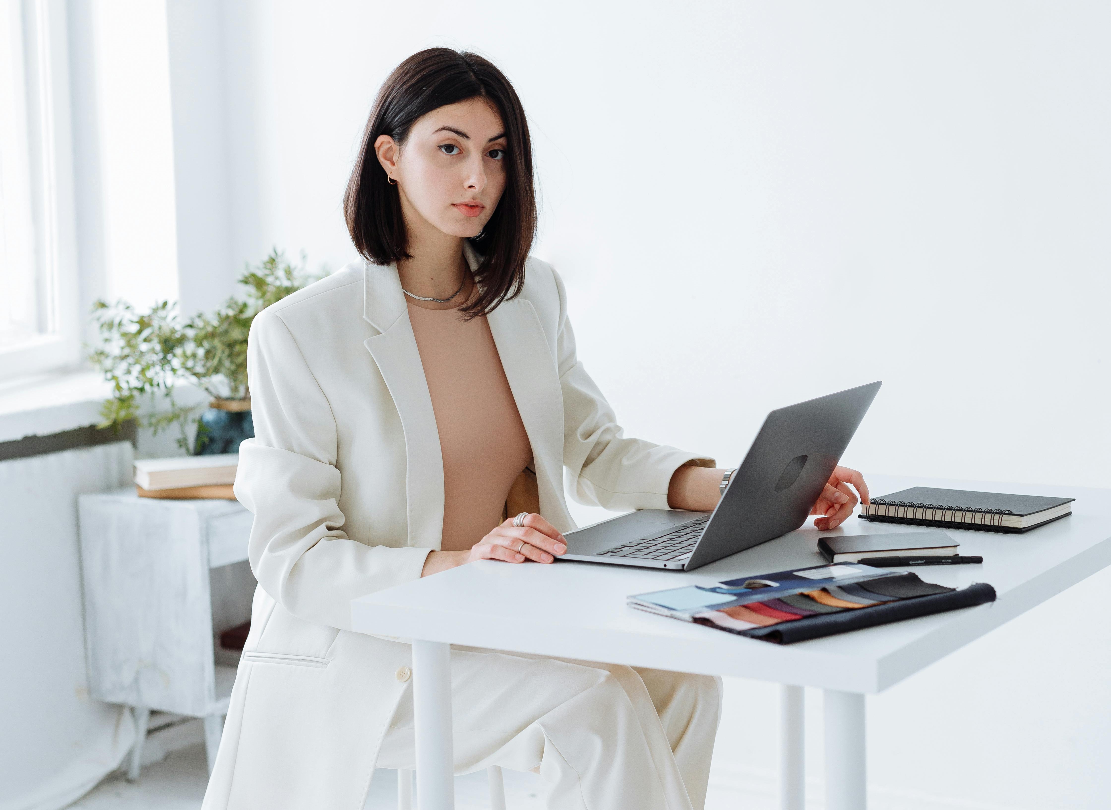 A young woman with dark, shoulder-length hair sits at a white desk in a bright, minimalist office. She is wearing an elegant cream-colored blazer and trousers over a beige top. She is looking directly at the camera while working on a laptop. On her desk, there are fabric color swatches and notebooks, suggesting a creative or fashion-related professional setting.