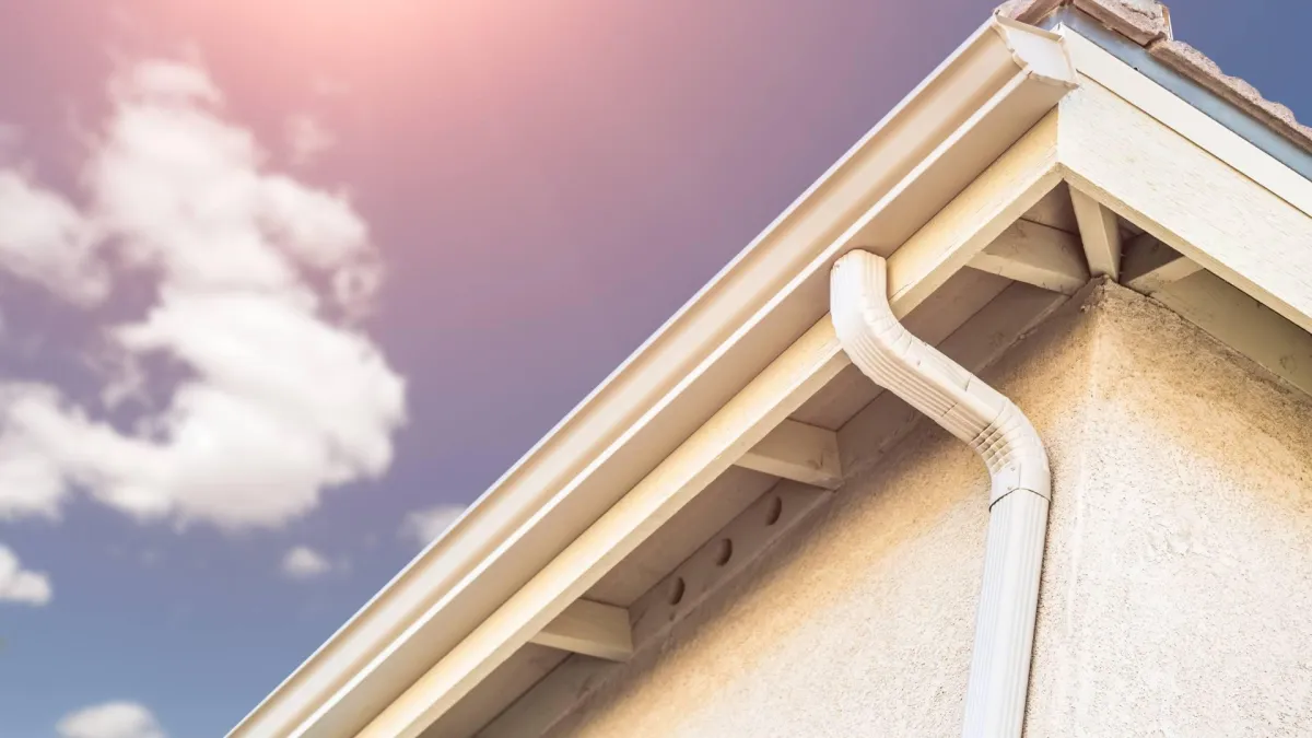 A home gutter is displayed on a cream colored home against a blue sky with white clouds.