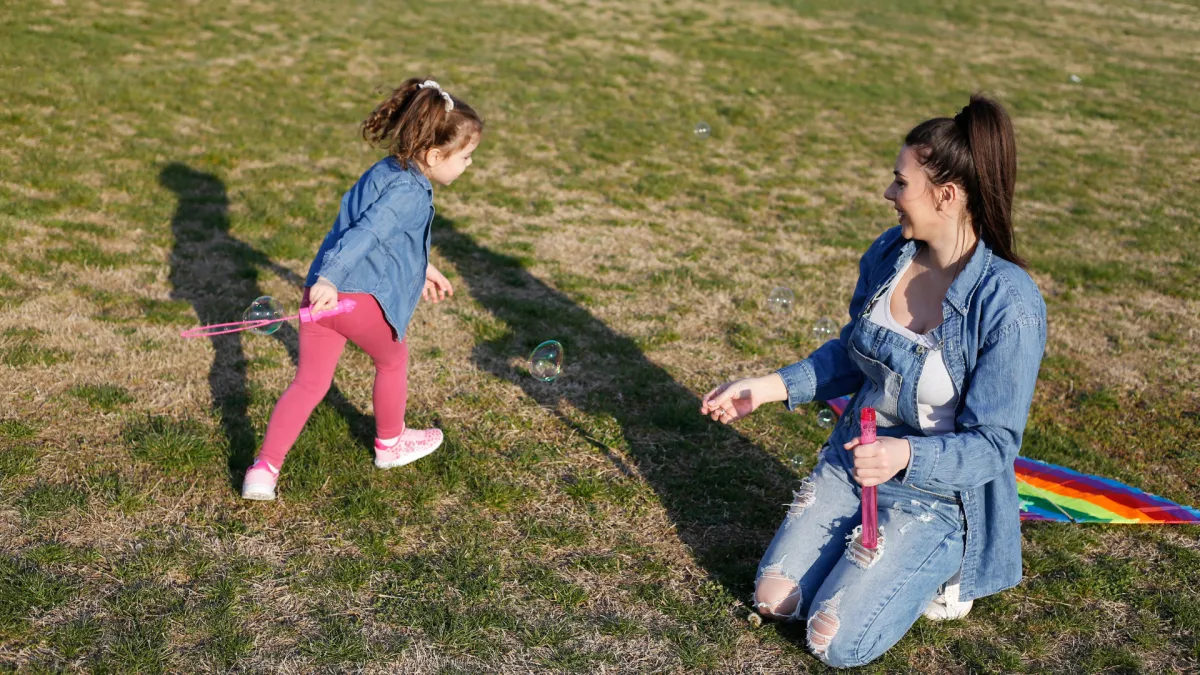 A mother kneels on the grass, smiling warmly at her young daughter who runs nearby with a bubble wand. They are outdoors on a sunny day, playing together and creating bubbles. Both wear denim outfits, and a colourful kite lies on the grass beside them. The scene captures joy, connection, and calm presence between parent and child.
