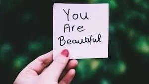 image of a woman's hand holding a post-it sticky note with the words "you are beautiful" written in black marker to represent the resiliency of women and importance of self care