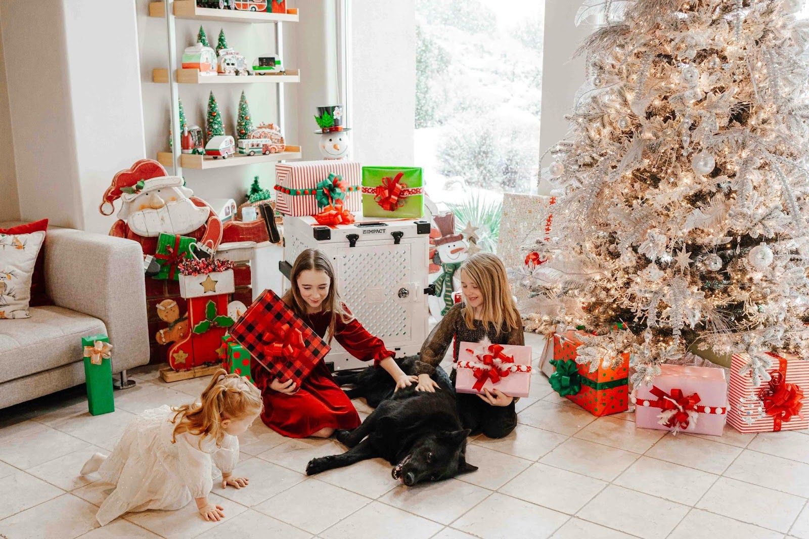 Two girls and a dog sit on the floor, smiling in front of a beautifully decorated Christmas tree.