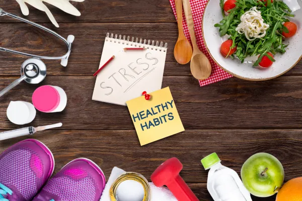 Flat lay of stress-related items including a notepad labeled stress, sticky note with healthy habits, workout gear, stethoscope, vegetables, fruits, and water bottle representing stress management and wellness.