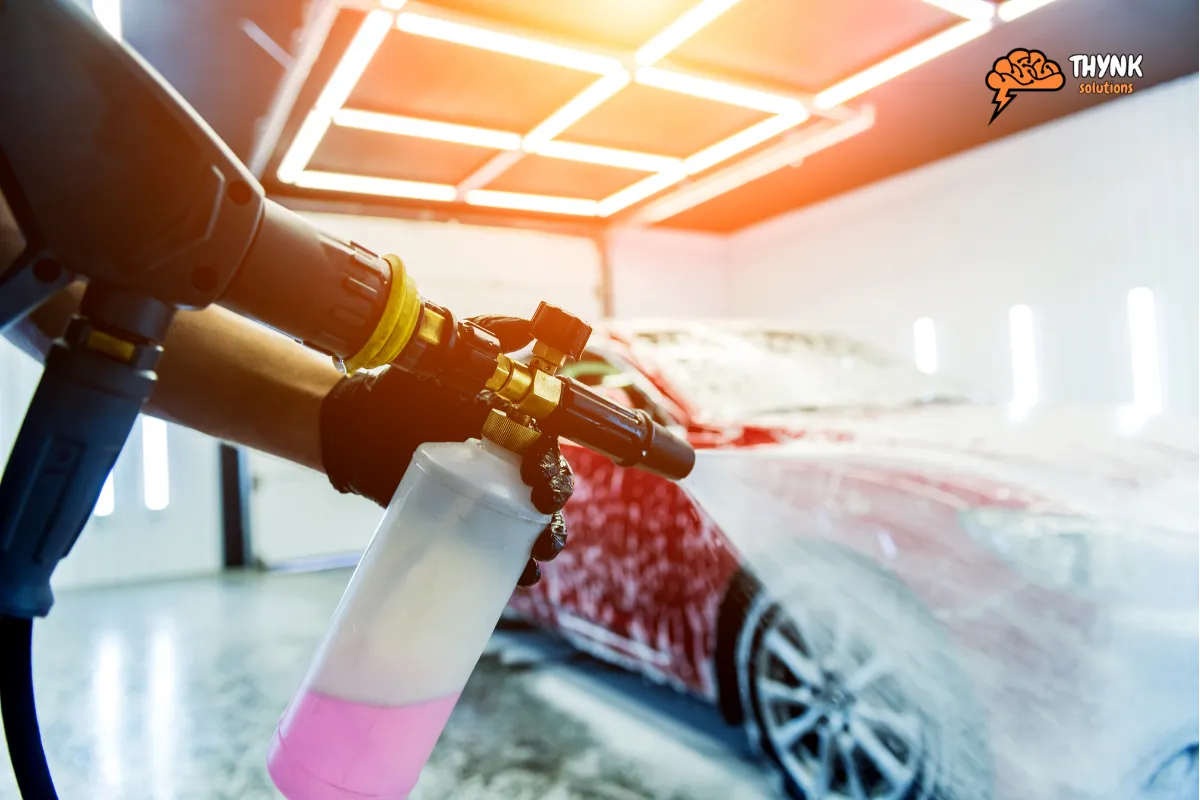 Auto detailer’s hand holding a soap spray gun in a garage setting with a car in the background.