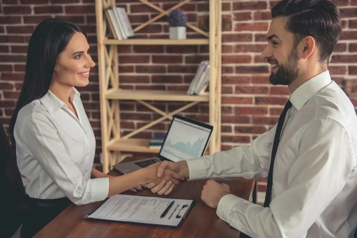 An attorney and client shake hands over a clipboard filled with paperwork.