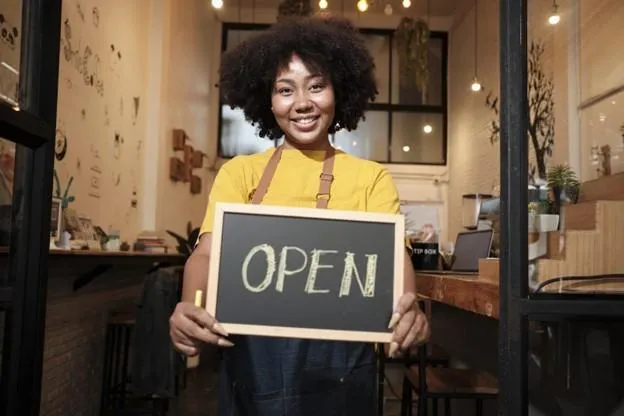Startup business owner in doorway of workshop, holding chalkboard sign that says, “OPEN” in pale yellow letters. 
