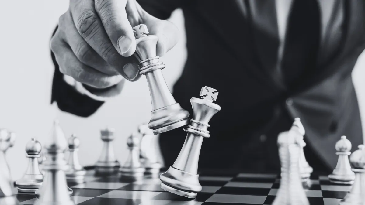 A hand, presumably that of a businessman in a formal suit, is captured holding a king chess piece while toppling another king on the chessboard.
