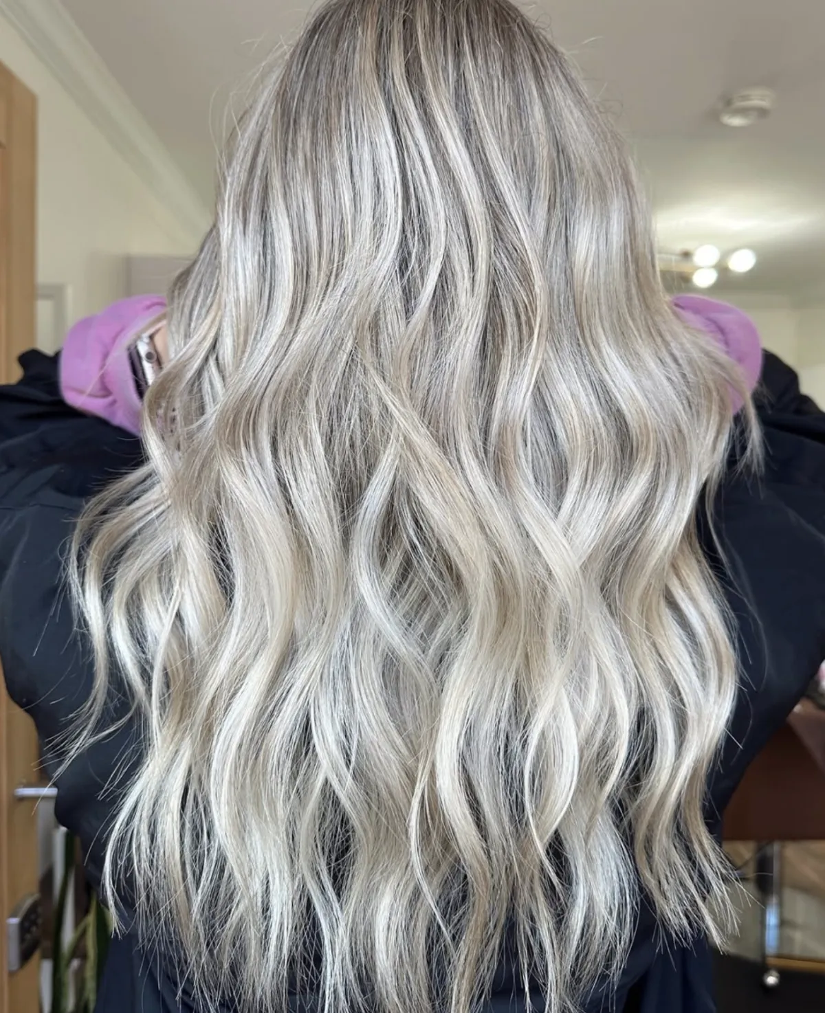Long cool blonde balayage styled in textured waves, showing blended highlights and dimensional tone in a salon setting.