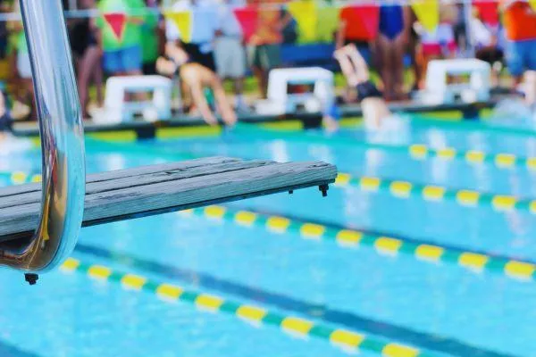 A diving board in sharp focus with a swim meet softly blurred in the background, capturing the anticipation and energy of the day