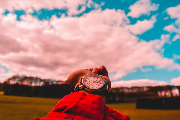 A close-up of an arm wearing a watch, pointing toward the vibrant colors of the sky and clouds, symbolizing the passage of time and the impact of daylight saving time.