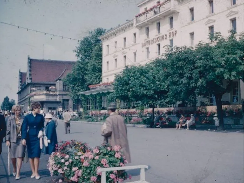 Vintage photo of everyday life in 1960s Germany, showing pedestrians on a sidewalk and a prominent building in the background
